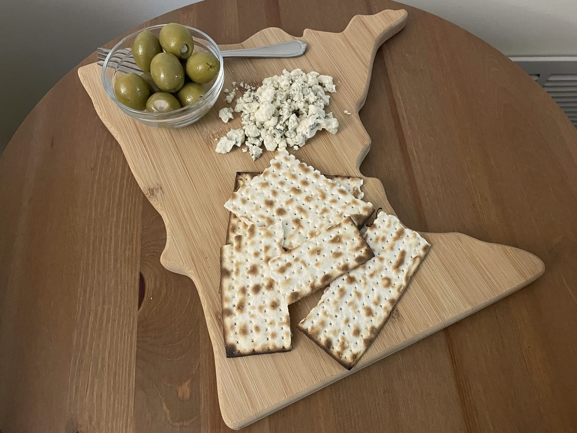 Serving board shaped like Minnesota with pieces of matzah, crumbled blue cheese, and a small bowl of olives on top