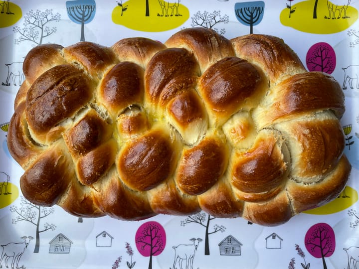 Six-stranded braided loaf of challah on a tray with pictures of cottages, goats, and trees